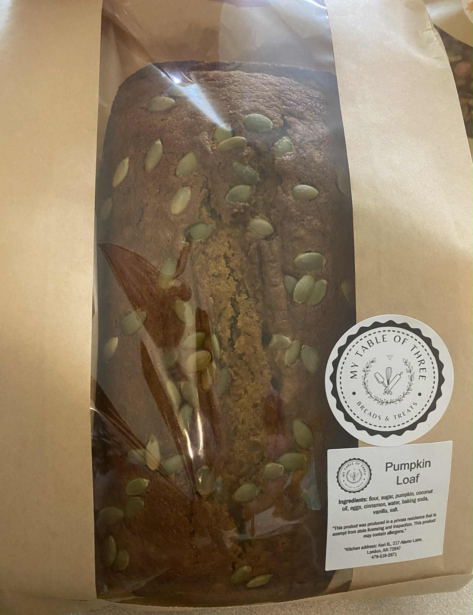 A pumpkin bread loaf with pumpkin seeds on top in a brown paper bag with My Table of Three Business labels on them.